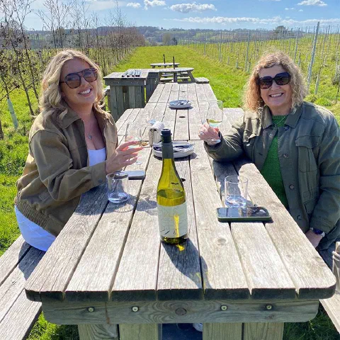 Two guests at a long wooden table with a bottle of white wine, vines behind them